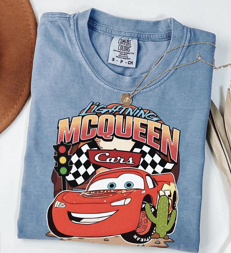 May include: A blue short-sleeve t-shirt featuring a graphic of the red race car Lightning McQueen from the movie Cars. The design includes the text Lightning McQueen, a traffic light, checkered flags, and a cactus. The shirt has a Comfort Colors label.