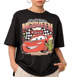 May include: A black short-sleeved t-shirt featuring a graphic print of the red character Lightning McQueen from the film Cars. The design includes the text Lightning McQueen, a traffic light, checkered flags, and a green cactus on a desert ground.