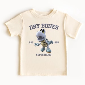 May include: Cream-colored t-shirt featuring a Dry Bones character from Super Mario. The design includes the text "DRY BONES" above the character, "EST 1985" and "SUPER MARIO" below. The graphic is in shades of gray, blue, and tan.