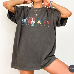 Może przedstawiać: Ciemnoszary t-shirt z kr&oacute;tkim rękawem z nadrukiem graficznym przedstawiającym Arielkę, Florka, Sebastiana oraz muszlę z perłą. Wz&oacute;r zawiera małe ż&oacute;łte gwiazdki i niebieskie kropki rozmieszczone na klatce piersiowej. Tkanina ma sprany wygląd.