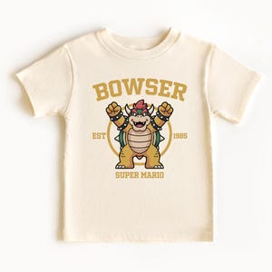 May include: Cream-colored t-shirt featuring a cartoon Bowser graphic from Super Mario. The design includes the word "BOWSER" in gold lettering above the character, with "EST 1985" and "SUPER MARIO" below.