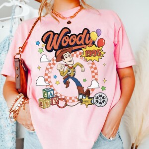 May include: A light pink t-shirt featuring a graphic print of the character Woody from Toy Story. The design includes the text Woody and 1995, colorful balloons, stars, alphabet blocks, a sheriff badge, and a film reel against a checkered circle.