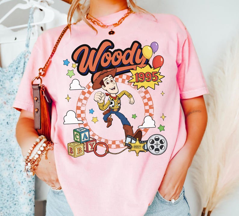 May include: A light pink t-shirt featuring a graphic print of the character Woody from Toy Story. The design includes the text Woody and 1995, colorful balloons, stars, alphabet blocks, a sheriff badge, and a film reel against a checkered circle.
