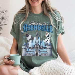 May include: A sage green short-sleeve t-shirt featuring a graphic of a blue ice castle, a reindeer, and a snowman. The text "Meet me in ARENDELLE" is printed in blue above the castle. The shirt has a crew neckline and a relaxed fit.