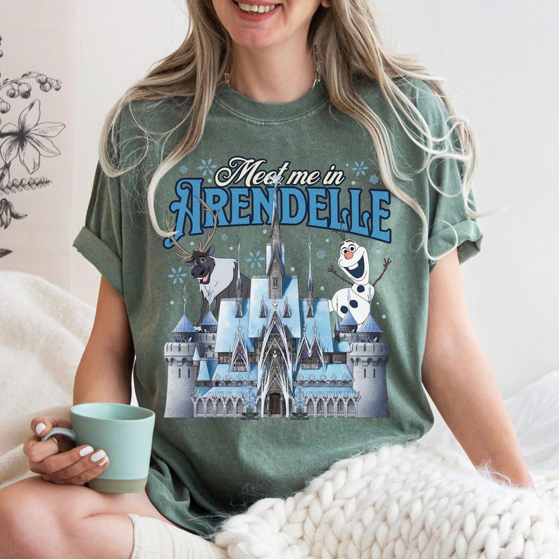 May include: A sage green short-sleeve t-shirt featuring a graphic of a blue ice castle, a reindeer, and a snowman. The text "Meet me in ARENDELLE" is printed in blue above the castle. The shirt has a crew neckline and a relaxed fit.