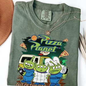 May include: An olive green crew neck t-shirt featuring a graphic print of three green three-eyed aliens. The design includes the text Pizza Planet and Out of This World Quality, along with illustrations of pizza slices and a yellow delivery truck.