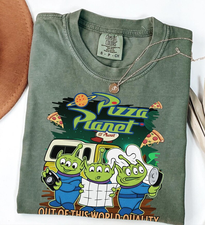 May include: An olive green crew neck t-shirt featuring a graphic print of three green three-eyed aliens. The design includes the text Pizza Planet and Out of This World Quality, along with illustrations of pizza slices and a yellow delivery truck.