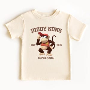 Könnte beinhalten: Cremefarbenes T-Shirt mit einem Diddy Kong-Motiv aus Super Mario. Das Design enthält den Text "DIDDY KONG", "EST 1985" und "SUPER MARIO". Der Cartoon-Affe trägt einen roten Hut und eine Weste. Ein lustiges, lässiges T-Shirt.