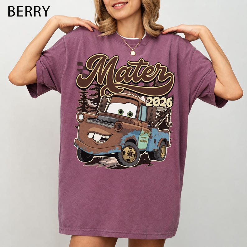 May include: A berry-colored short-sleeve t-shirt featuring a graphic print of the character Mater from the movie Cars. The design includes the text Mater and 2026 in a brown script font, set against a background of dark forest silhouettes.