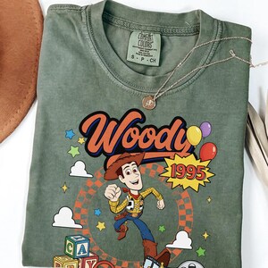 May include: An olive green t-shirt featuring a graphic of the character Woody from Toy Story. The design includes the text Woody, the year 1995, colorful stars, clouds, balloons, alphabet blocks, and a film reel. The shirt has a Comfort Colors label.