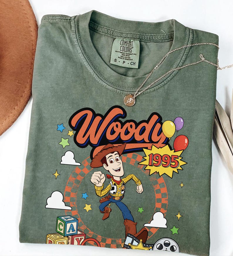 May include: An olive green t-shirt featuring a graphic of the character Woody from Toy Story. The design includes the text Woody, the year 1995, colorful stars, clouds, balloons, alphabet blocks, and a film reel. The shirt has a Comfort Colors label.
