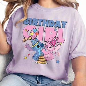May include: A light purple t-shirt featuring a graphic of the characters Stitch and Angel from Lilo & Stitch. The text "BIRTHDAY GIRL" is in blue and pink. The characters are celebrating with a cake and party hats.