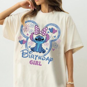 May include: Cream-colored t-shirt featuring a blue cartoon character with a purple bow and ears, surrounded by blue Mickey Mouse-shaped designs with fireworks and balloons. The text "Birthday Girl" is written in blue and pink.