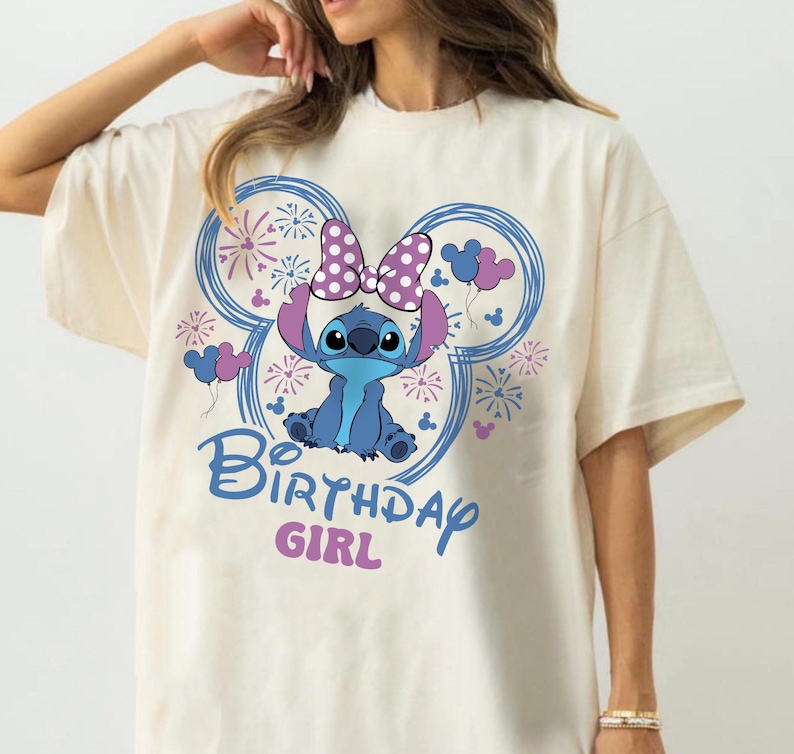 May include: Cream-colored t-shirt featuring a blue cartoon character with a purple bow and ears, surrounded by blue Mickey Mouse-shaped designs with fireworks and balloons. The text "Birthday Girl" is written in blue and pink.