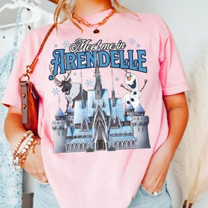 May include: A light pink short-sleeve t-shirt featuring a graphic print of a blue ice castle, a reindeer, and a snowman. The text "Meet me in ARENDELLE" is printed in blue above the castle, surrounded by small blue snowflake illustrations.