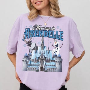 May include: A light purple short-sleeve t-shirt featuring a graphic print of an ice castle, a reindeer, and a snowman. The text "Meet me in ARENDELLE" is printed in blue and black above the castle, surrounded by small blue snowflake icons.