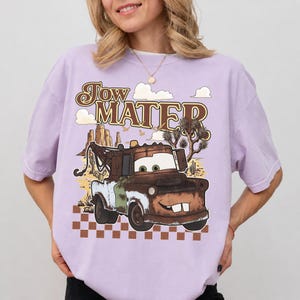 May include: A light purple t-shirt featuring a graphic of a rusty brown tow truck character with large eyes and two front teeth. The text "Tow Mater" appears above the truck, set against a desert landscape with cacti and a checkered pattern.