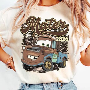 May include: A cream-colored t-shirt featuring a graphic print of a brown and blue tow truck character with large eyes and buck teeth. The text "Mater" appears in large brown script above the truck, with the year "2026" printed to the right.