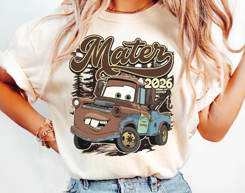May include: A cream-colored t-shirt featuring a graphic print of a brown and blue tow truck character with large eyes and buck teeth. The text "Mater" appears in large brown script above the truck, with the year "2026" printed to the right.