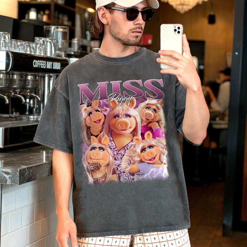 May include: Dark gray t-shirt featuring a graphic of Miss Piggy from The Muppets. The design includes the text "MISS Piggy" in a pink and purple font, along with multiple images of Miss Piggy. The shirt has a vintage, worn look.