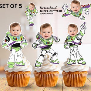 May include: Set of 5 personalised Buzz Lightyear cupcake toppers. Each cupcake features a smiling baby's face in a Buzz Lightyear costume. The cupcakes have white frosting and a golden-brown cake base.