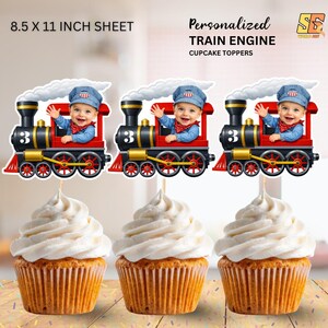 May include: Three cupcakes topped with white frosting, each adorned with a personalized train engine cupcake topper. The toppers feature a smiling baby in a conductor's outfit, waving from a red, black, and gold train.