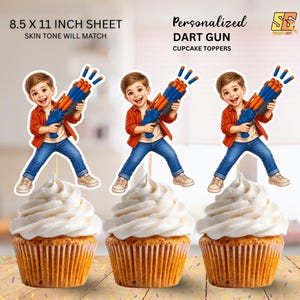 May include: Cupcake toppers featuring a cartoon boy holding a blue and orange dart gun. The toppers are placed on top of cupcakes with white frosting. The text "Personalized DART GUN CUPCAKE TOPPERS" is visible.