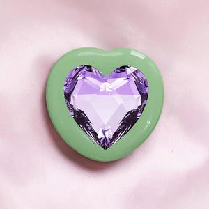 Puede incluir: Un dije en forma de corazón con un borde liso de color verde claro que rodea un corazón de cristal violeta facetado de talla brillante en el centro. El objeto tiene un acabado brillante y una apariencia estratificada con una superficie reflectante.