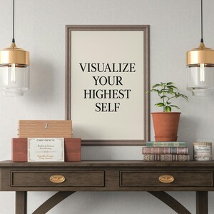 May include: A framed print with the text "VISUALIZE YOUR HIGHEST SELF" sits on a dark wood table. The table holds a plant in a terracotta pot, books, and decorative boxes. Two gold and glass pendant lights hang above.