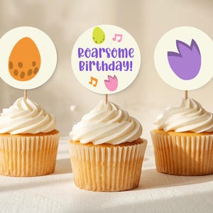 Puede incluir: Tres cupcakes con glaseado blanco y adornos decorativos. Uno tiene un diseño de huevo naranja, otro dice "Roarsome Birthday!", y el tercero tiene un diseño de tulipán morado. Los cupcakes están en moldes de papel acanalados.