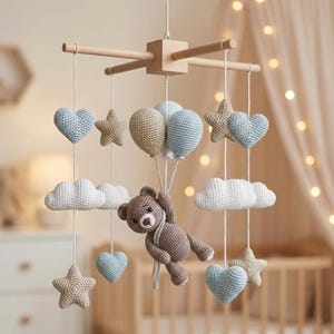 May include: A handmade wooden baby mobile featuring crocheted elements. The mobile includes a brown teddy bear, blue and tan hearts, stars, clouds, and balloons. The nursery decor is suspended from a wooden frame.