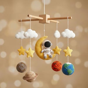 Könnte beinhalten: Handgefertigtes Baby-Mobile mit einem gehäkelten Astronauten auf einem gelben Halbmond. Es zeigt weiße Wolken, gelbe Sterne und Planetenkugeln, die alle an einem Holzrahmen hängen. Die Farbgebung ist weich und einladend.