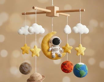 May include: A handmade baby mobile featuring a crocheted astronaut seated on a yellow crescent moon. The mobile includes white clouds, yellow stars, and planet-themed spheres, all suspended from a wooden frame. The color palette is soft and inviting.