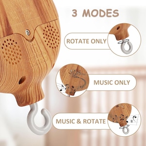 Op de afbeelding: Een babymobielmotor met een houtnerf-afwerking, voorzien van een witte haak en luidsprekerroosters. De tekst 3 MODES wordt getoond met drie cirkelvormige diagrammen gelabeld ROTATE ONLY, MUSIC ONLY en MUSIC & ROTATE voor de instellingen.