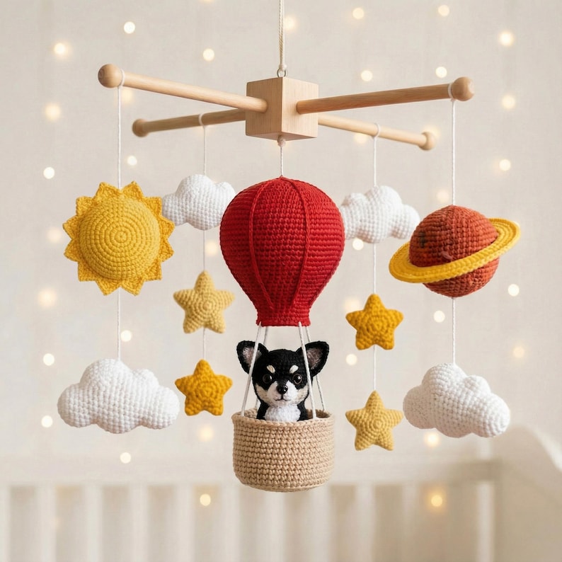 Op de afbeelding: Een gehaakte babymobiel met een centrale rode heteluchtballon die een zwart-witte hond in een beige mandje draagt. Omringende elementen zijn een gele zon, een planeet met ring, witte wolken en gele sterren opgehangen aan een houten frame.