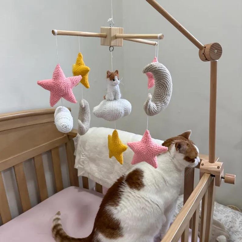 Op de afbeelding: Een houten wiegmobiel met gehaakte hangende ornamenten, waaronder roze en gele sterren, een beige halve maan, een witte wolk en een klein gehaakt kattenfiguurtje dat op een wolk zit, opgehangen aan een centraal houten kruisframe.