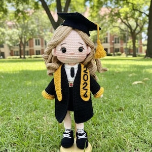 Puede incluir: Muñeca de ganchillo con un vestido de graduación negro con ribetes dorados y un birrete a juego. La muñeca tiene el pelo rubio y está de pie sobre una base de madera. El año "2022" aparece en el vestido.
