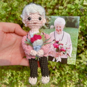 May include: A crocheted doll of a woman with white hair, wearing a pink cardigan and black trousers, holding a bouquet of flowers. A photograph of a woman holding flowers is next to the doll. The doll has pearl earrings and a pearl necklace.
