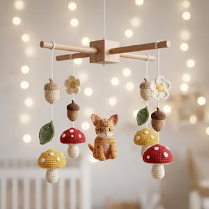 Op de afbeelding: Een hangende mobiel met een centraal houten kruisframe waaraan witte koorden gehaakte figuren vasthouden, waaronder een rosse kat, rode en gele paddenstoelen met witte stippen, groene bladeren, beige eikels en witte bloemen met gele harten.