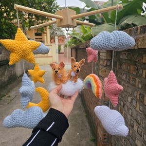 Op de afbeelding: Een hangende mobiel met gehaakte elementen aan een houten frame. De mobiel bevat gele sterren, een gele maansikkel, lichtblauwe en witte wolken, roze sterren, een kleine regenboog en twee bruine gehaakte honden op een witte wolk.