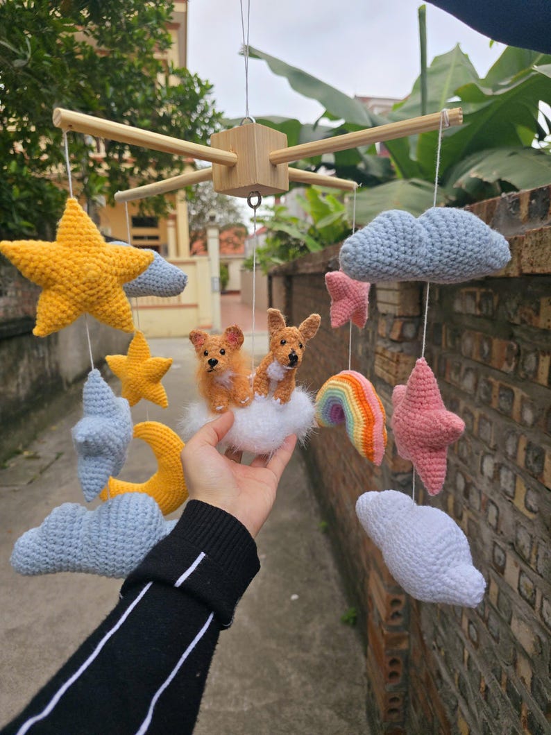 Op de afbeelding: Een hangende mobiel met gehaakte elementen aan een houten frame. De mobiel bevat gele sterren, een gele maansikkel, lichtblauwe en witte wolken, roze sterren, een kleine regenboog en twee bruine gehaakte honden op een witte wolk.