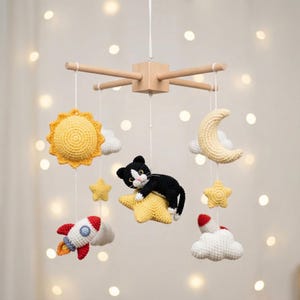 Op de afbeelding: Een hangende mobiel met gehaakte figuren aan een houten kruisframe. De figuren bestaan uit een zwart-witte kat op een gele ster, een gele zon, een gele halve maan, witte wolken, gele sterren en rood-witte raketten.