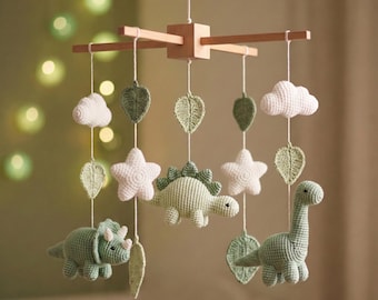 May include: A hanging mobile featuring crocheted figures suspended from a wooden cross frame. The mobile includes two green dinosaurs, two white stars, two white clouds, and several green leaves, all crafted with a textured yarn finish.