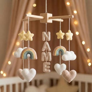 Pode incluir: Móvel de bebê artesanal com estrelas, nuvens, arco-íris e corações de crochê em tons pastéis suaves. O móvel inclui as letras "NAME" e é suspenso por uma estrutura de madeira, criando uma decoração lúdica para o berçário.