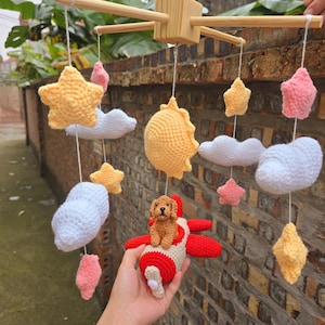 Op de afbeelding: Een hangende mobiel met gehaakte elementen, waaronder een bruine hond in een rood-wit vliegtuig, een gele zon, witte wolken en roze en gele sterren, opgehangen aan een lichtgekleurd houten kruisvormig frame met witte koordjes.