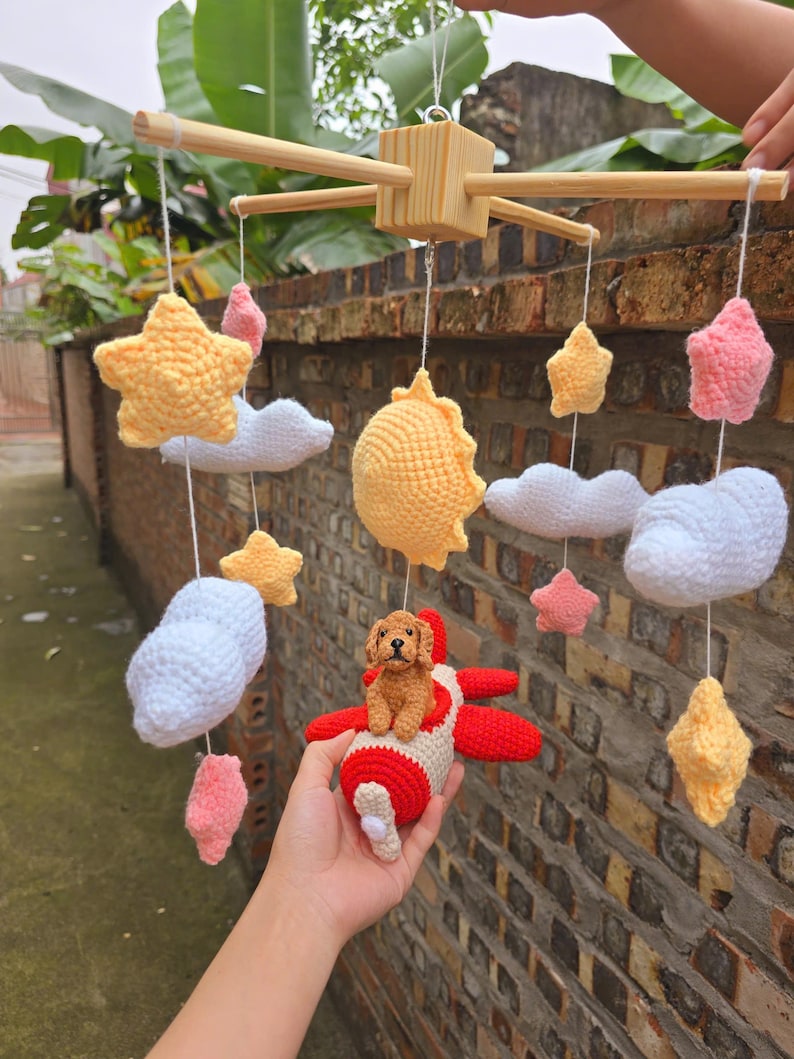 Op de afbeelding: Een hangende mobiel met gehaakte elementen, waaronder een bruine hond in een rood-wit vliegtuig, een gele zon, witte wolken en roze en gele sterren, opgehangen aan een lichtgekleurd houten kruisvormig frame met witte koordjes.