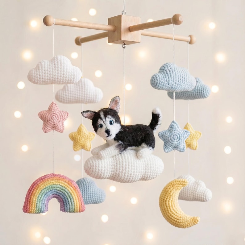 Op de afbeelding: Een hangende mobiel met een centrale gehaakte husky-hond die op een witte wolk rust. De mobiel bevat gehaakte elementen zoals witte en blauwe wolken, gele en roze sterren, een gele halve maan en een regenboog in pastelkleuren.