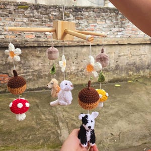 Op de afbeelding: Een hangende mobiel met een houten kruisframe en bungelende gehaakte figuren, waaronder een rood-witte paddenstoel, een bruine eikel, een witte bloem, een beige hond, een witte hond, een zwart-witte hond en een gele paddenstoel.