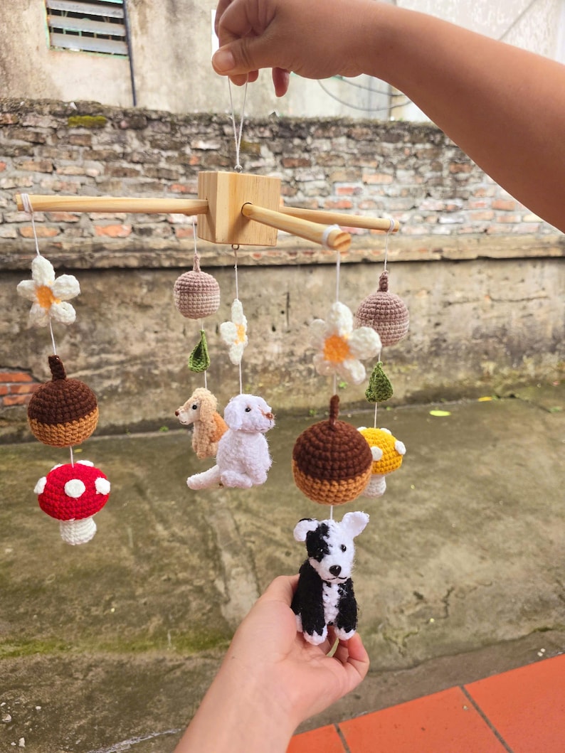 Op de afbeelding: Een hangende mobiel met een houten kruisframe en bungelende gehaakte figuren, waaronder een rood-witte paddenstoel, een bruine eikel, een witte bloem, een beige hond, een witte hond, een zwart-witte hond en een gele paddenstoel.