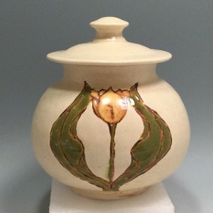 May include: Cream-colored ceramic jar with a lid, featuring a hand-painted tulip design in shades of yellow, green, and brown. The jar has a rounded shape and a matching lid. A decorative piece for the home.